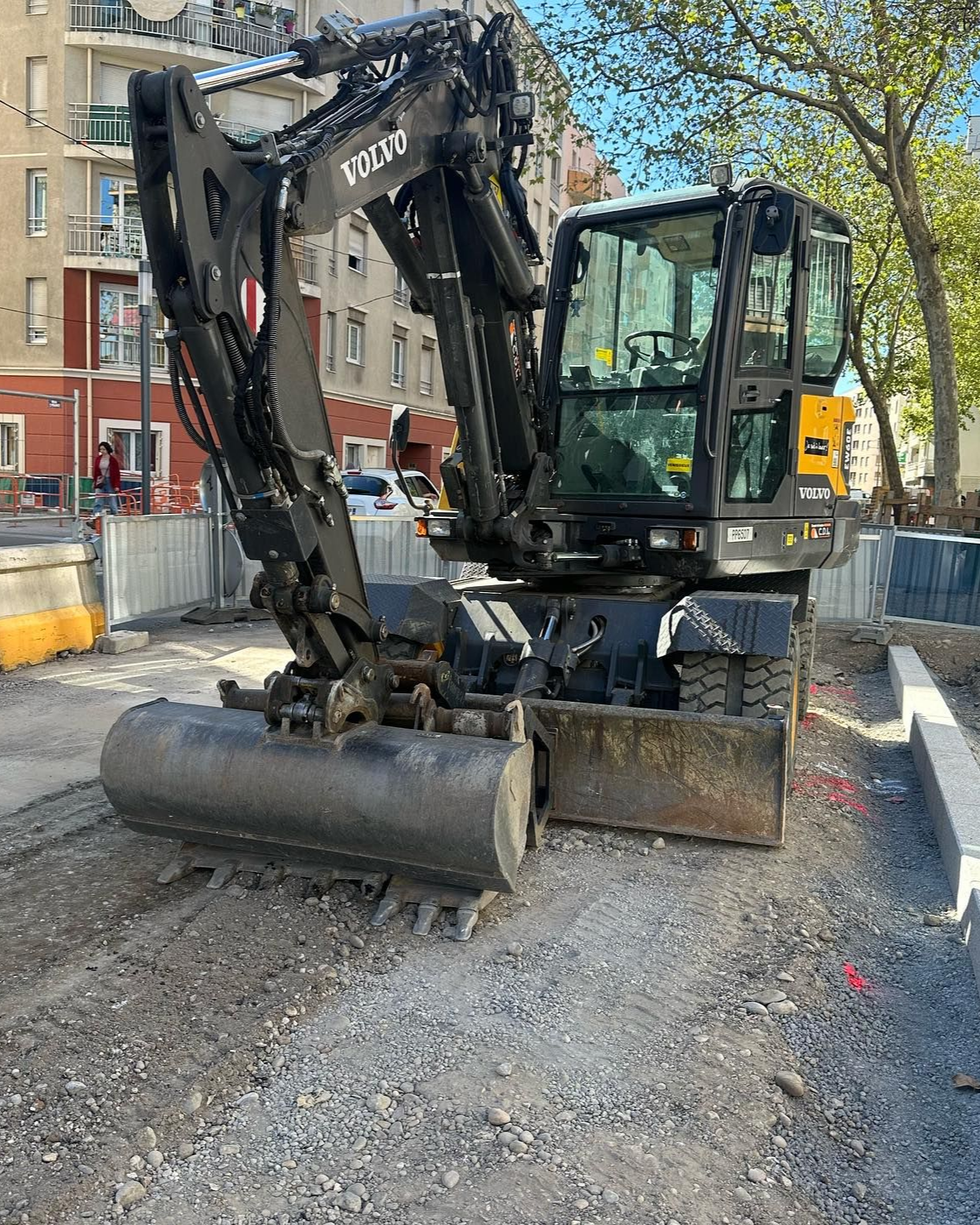 Une pelleteuse noire avec lame se trouve dans une rue recouverte de gravier, à côté d'immeubles et d'arbres.