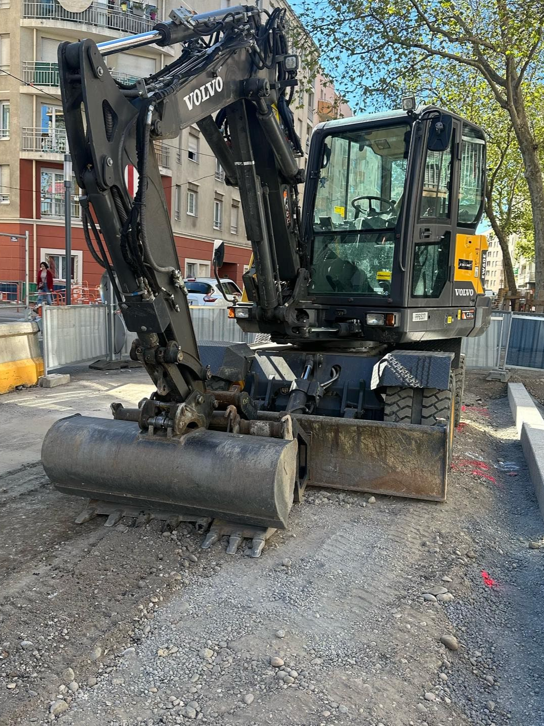 Une pelleteuse noire est stationnée sur un chantier de voirie, entourée de barrières, de gravier, d'arbres et d'immeubles d'habitation.