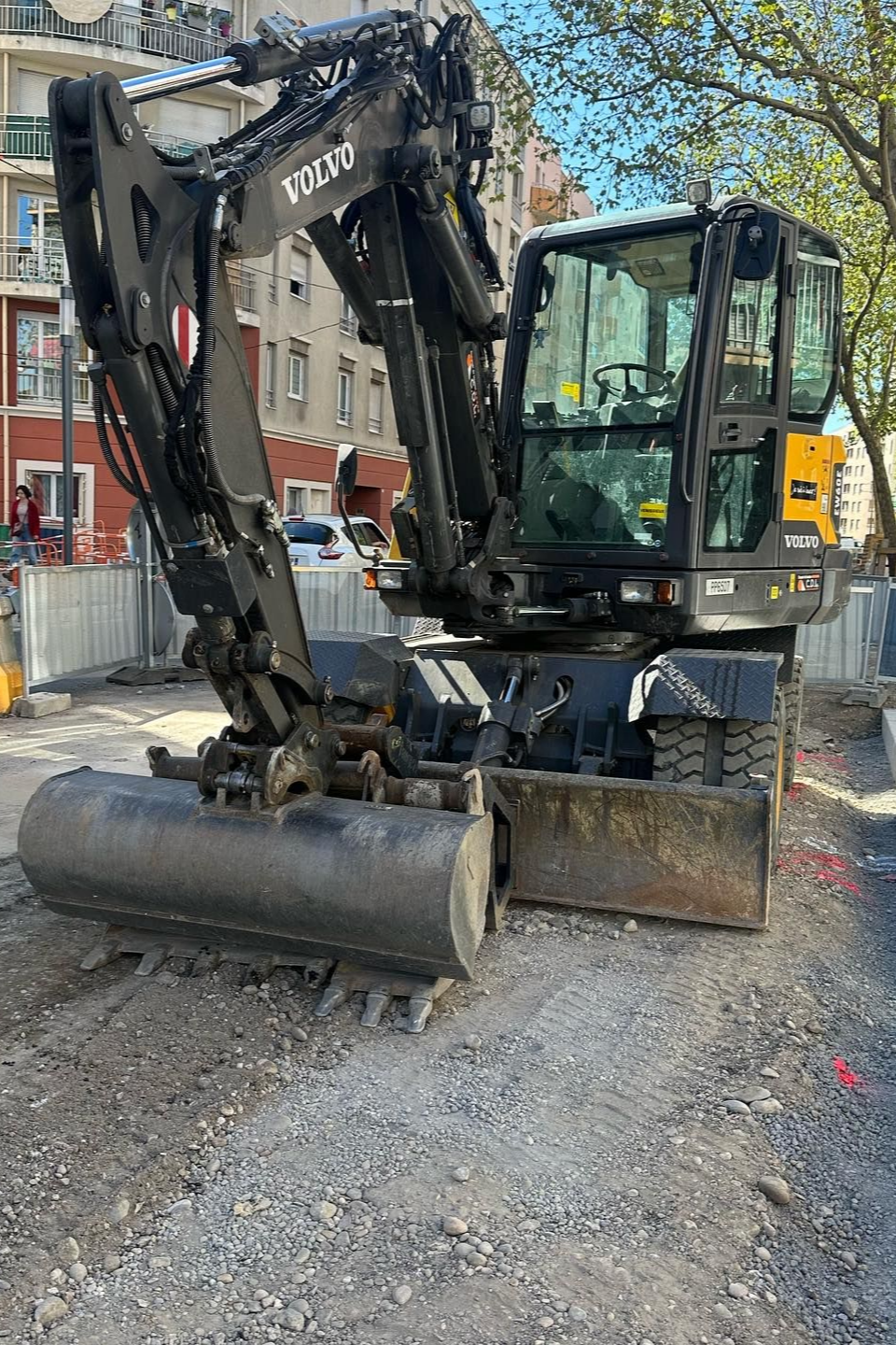 Une pelleteuse noire avec son godet est garée sur une rue de gravier, à côté de bâtiments et d'arbres.