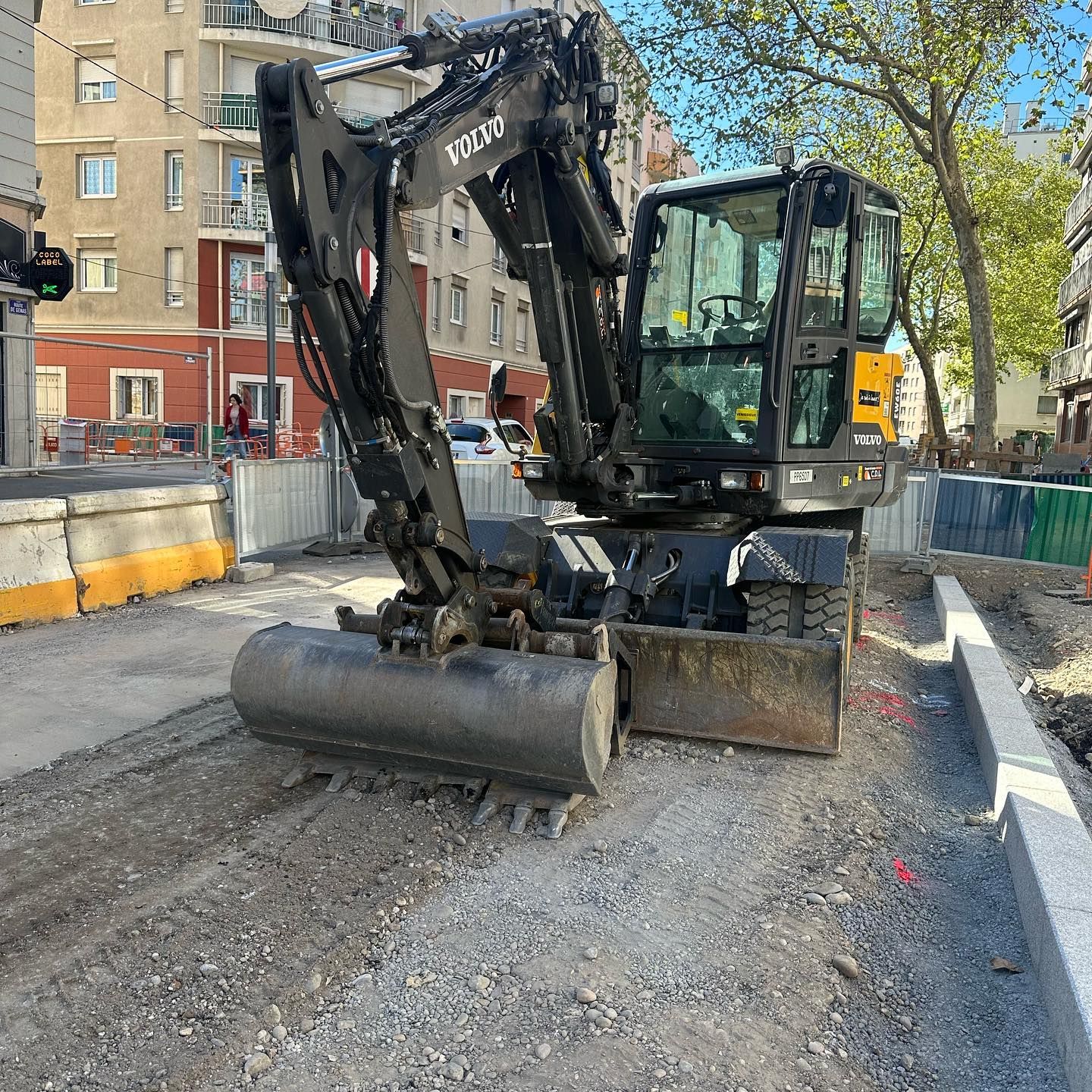 Une excavatrice compacte du gravier sur un chantier de construction dans une rue, à proximité de bâtiments et d'arbres.