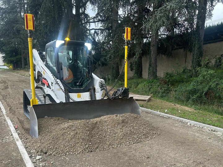 Chargeuse compacte blanche poussant du gravier sur un chantier routier, avec des poteaux de guidage jaunes levés.