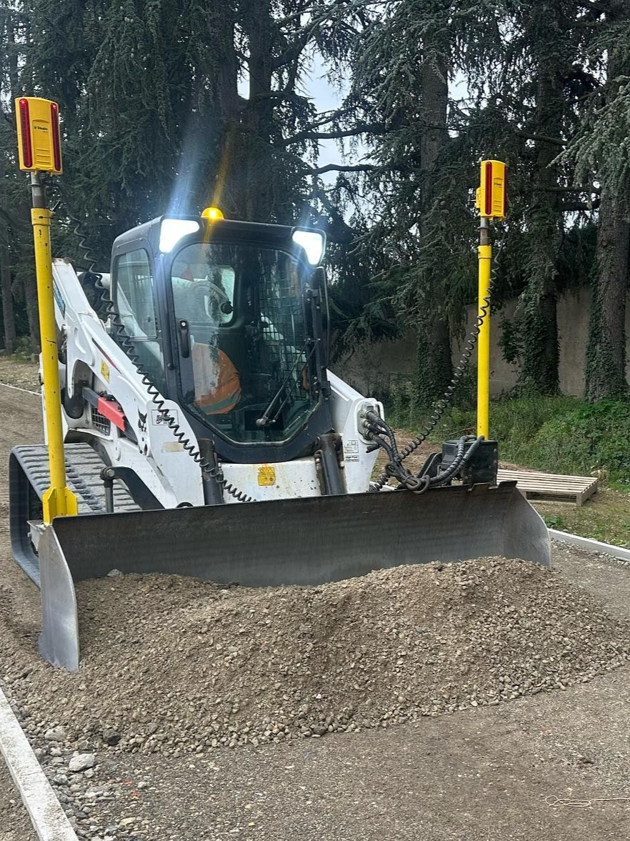 Une chargeuse compacte pousse du gravier avec son godet sur un chemin de terre bordé d'arbres.