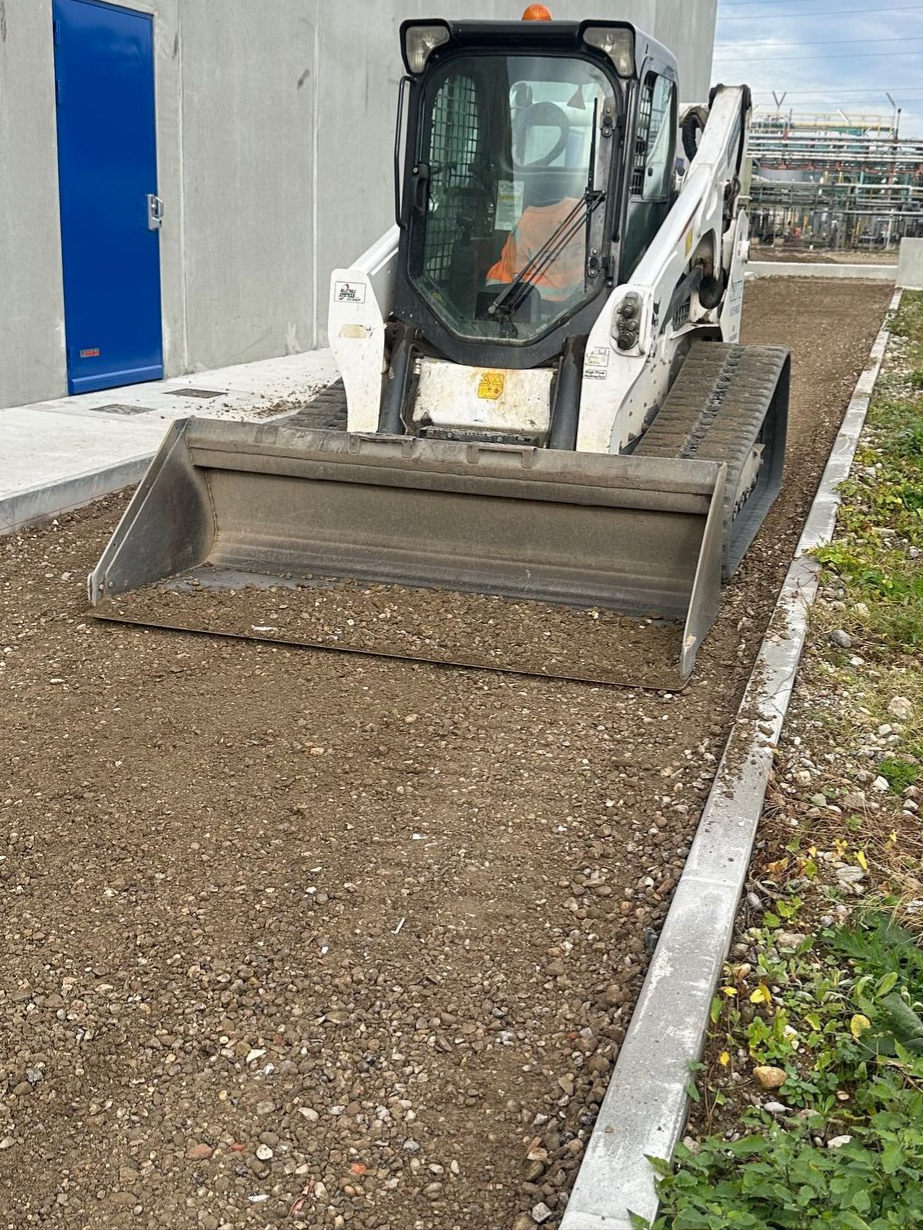 Chargeuse compacte blanche avec godet sur un chemin de gravier à côté d'un bâtiment.