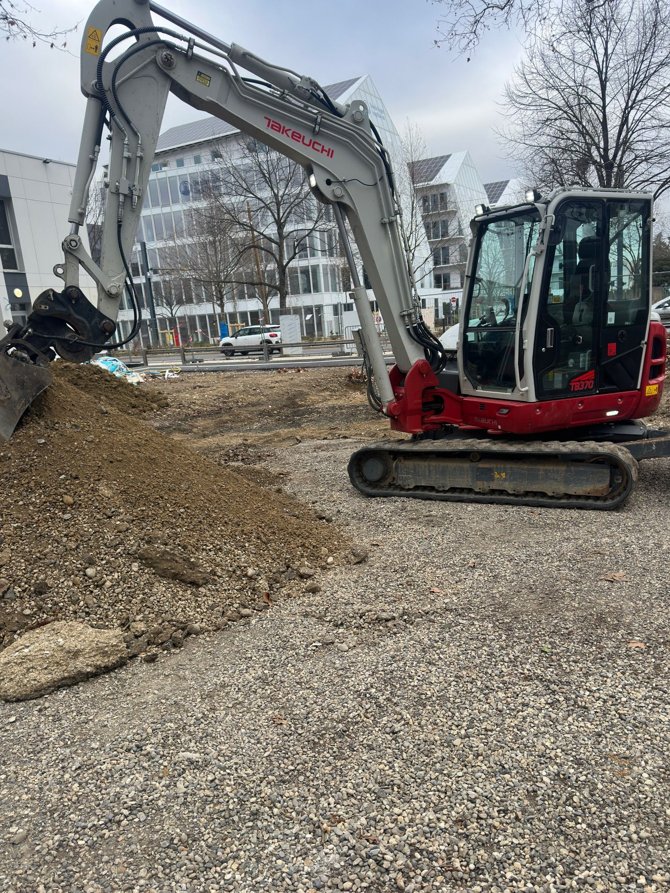 Une excavatrice sur un chantier de construction en gravier, à côté d'un trottoir clôturé et de bâtiments en briques.