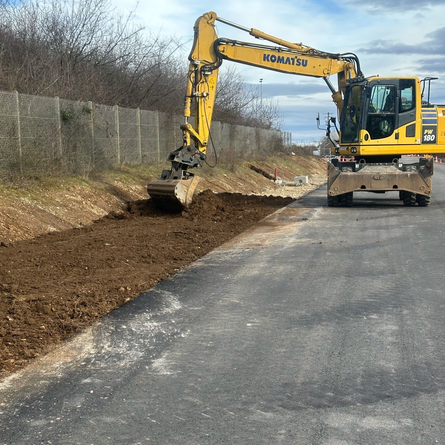 Une pelleteuse jaune nivelle le sol au bord d'une route goudronnée, avec une clôture et des arbres dénudés en arrière-plan.