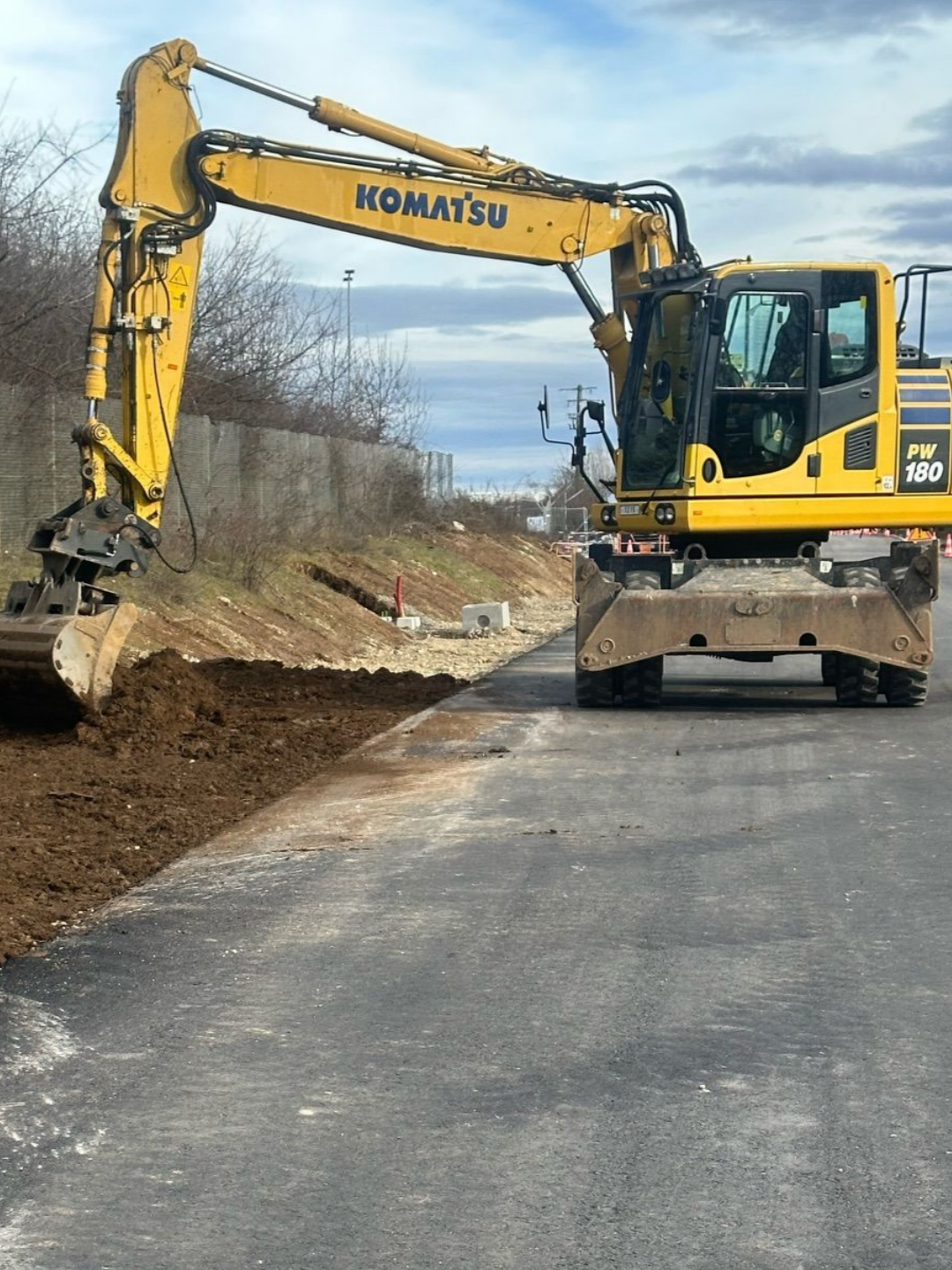 Une pelle mécanique Komatsu jaune en bordure d'une route goudronnée, creusant la terre sous un ciel nuageux.