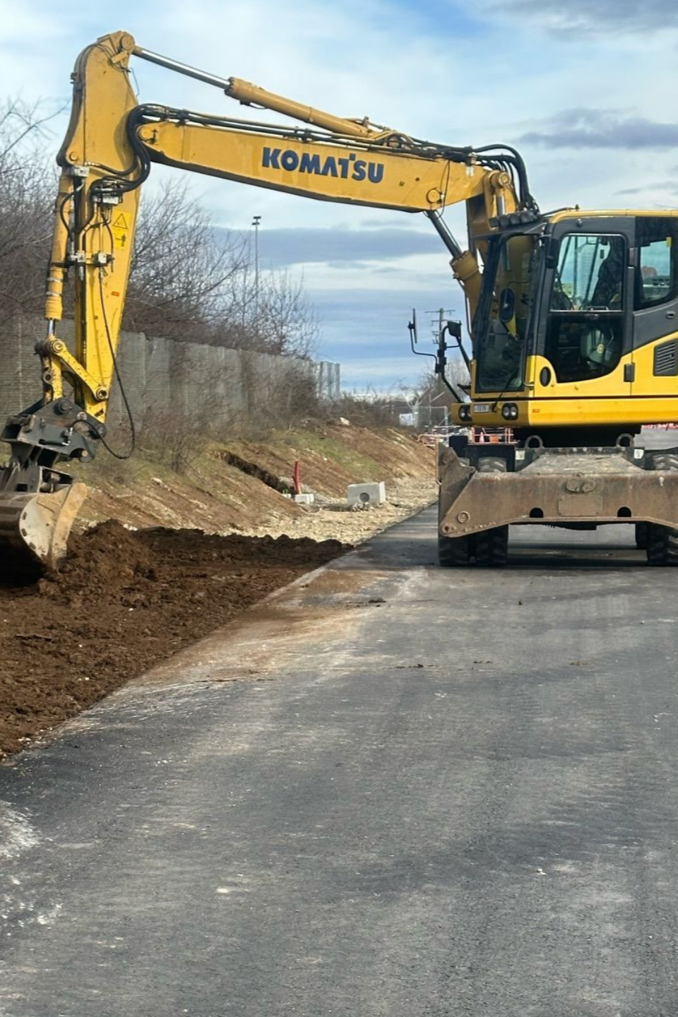 Une pelle mécanique Komatsu jaune en bordure d'une route goudronnée, creusant la terre sous un ciel nuageux.
