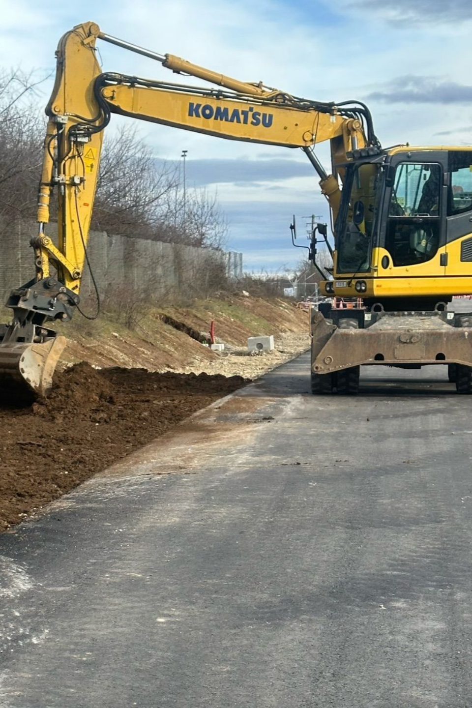 Une pelle Komatsu jaune creuse en bord de route sous un ciel nuageux.