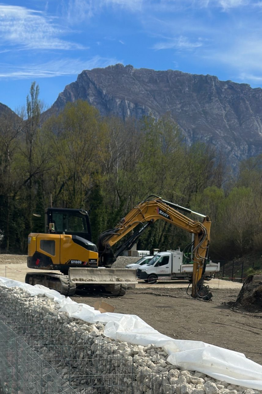 Une pelleteuse jaune et une camionnette blanche sur un terrain boueux, avec des montagnes et un ciel bleu en arrière-plan.