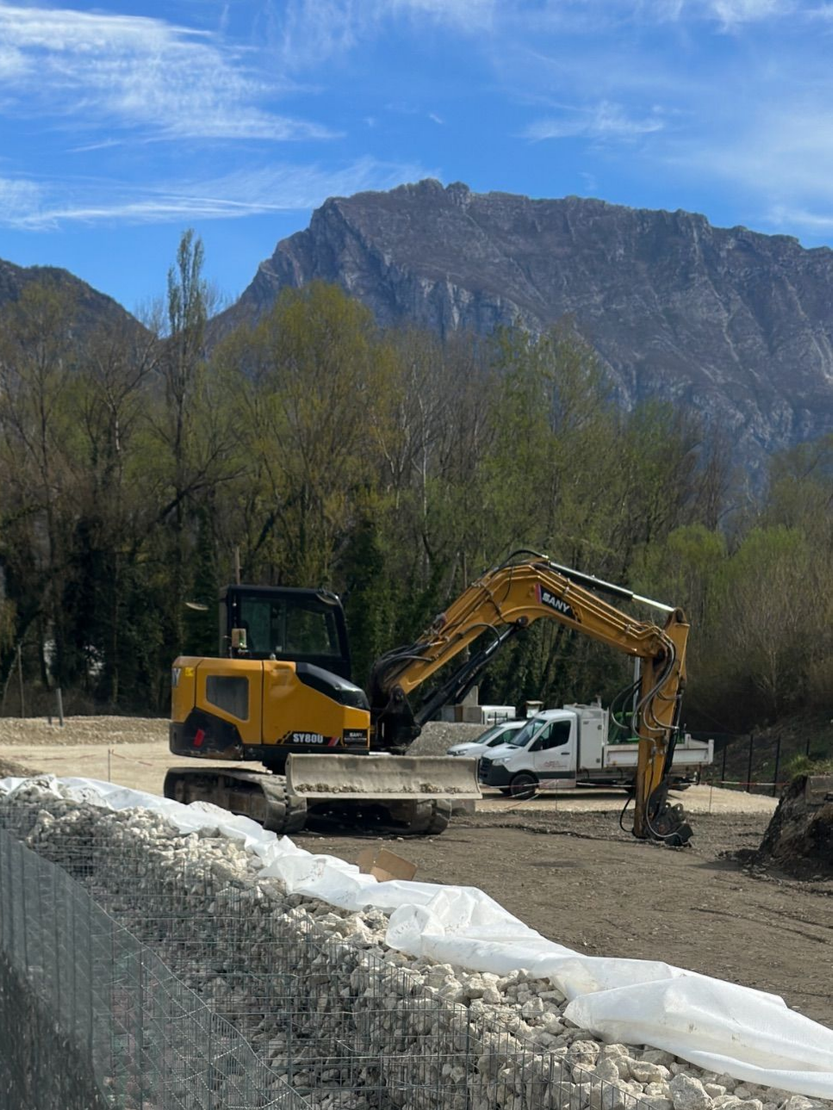 Chantier de construction avec une pelleteuse jaune et un camion blanc au milieu du gravier, avec des montagnes en arrière-plan.