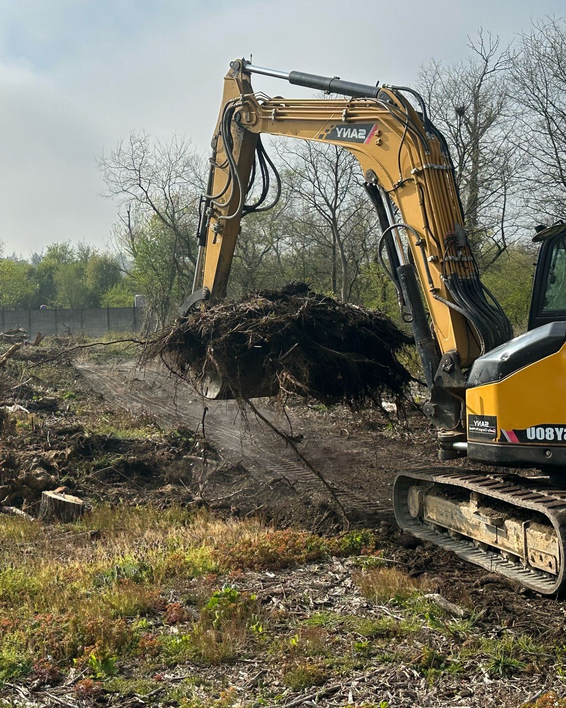 Une excavatrice jaune soulève un gros tas de terre sur un chantier extérieur boueux.