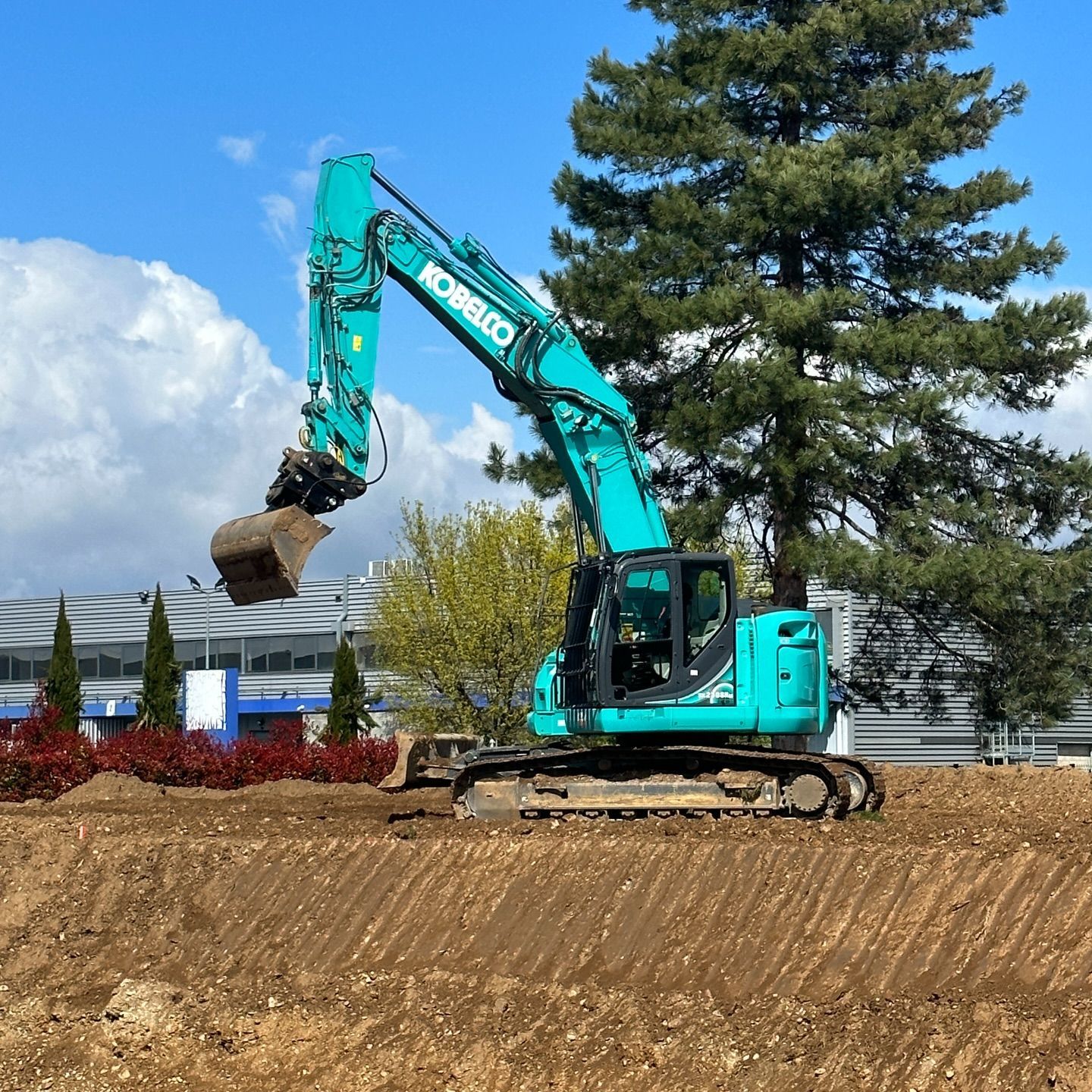 Une excavatrice turquoise soulève de la terre sur un chantier de construction bordé d'arbres et d'aménagements paysagers.