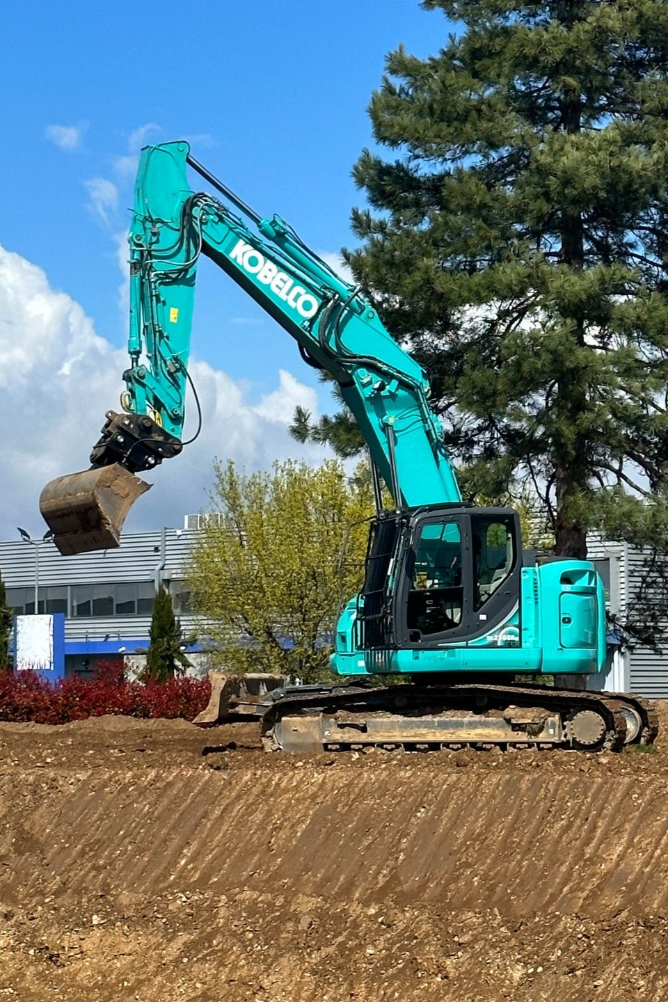 Une pelleteuse turquoise soulève de la terre sur un chantier de construction à côté d'arbres et d'une maison.