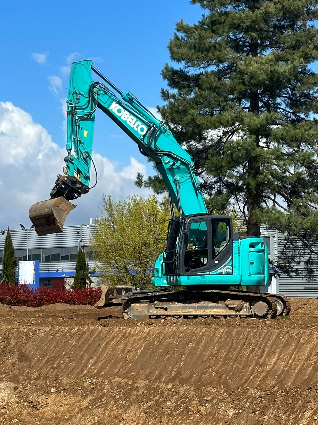 Une pelleteuse turquoise creuse un terrain vague sous un ciel bleu azur, avec des arbres et des maisons en arrière-plan.