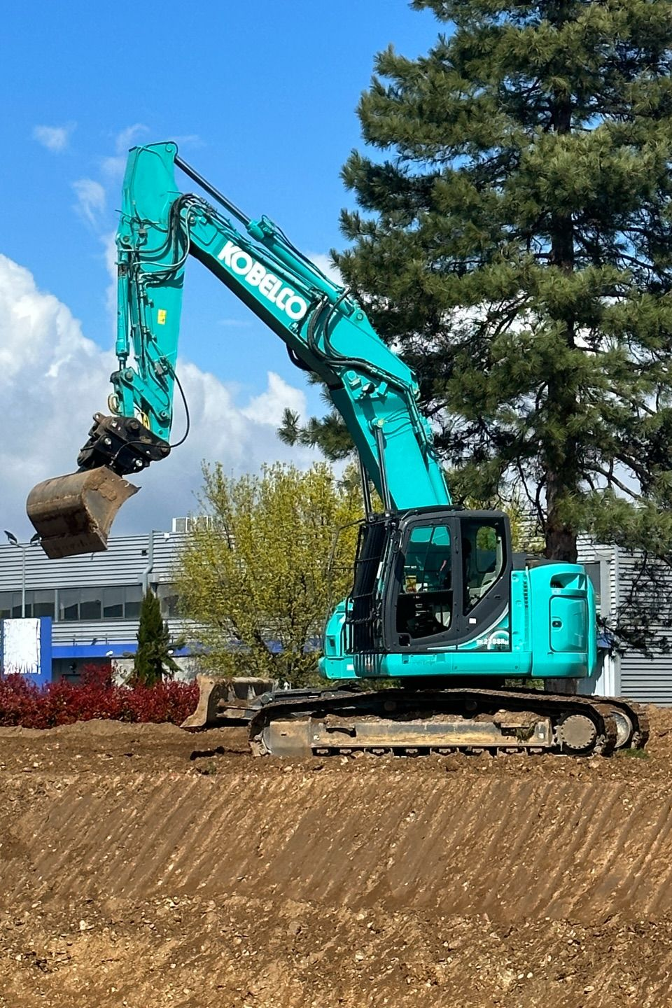 Une excavatrice turquoise sur un terrain en terre battue, à côté d'arbres et d'un bâtiment.