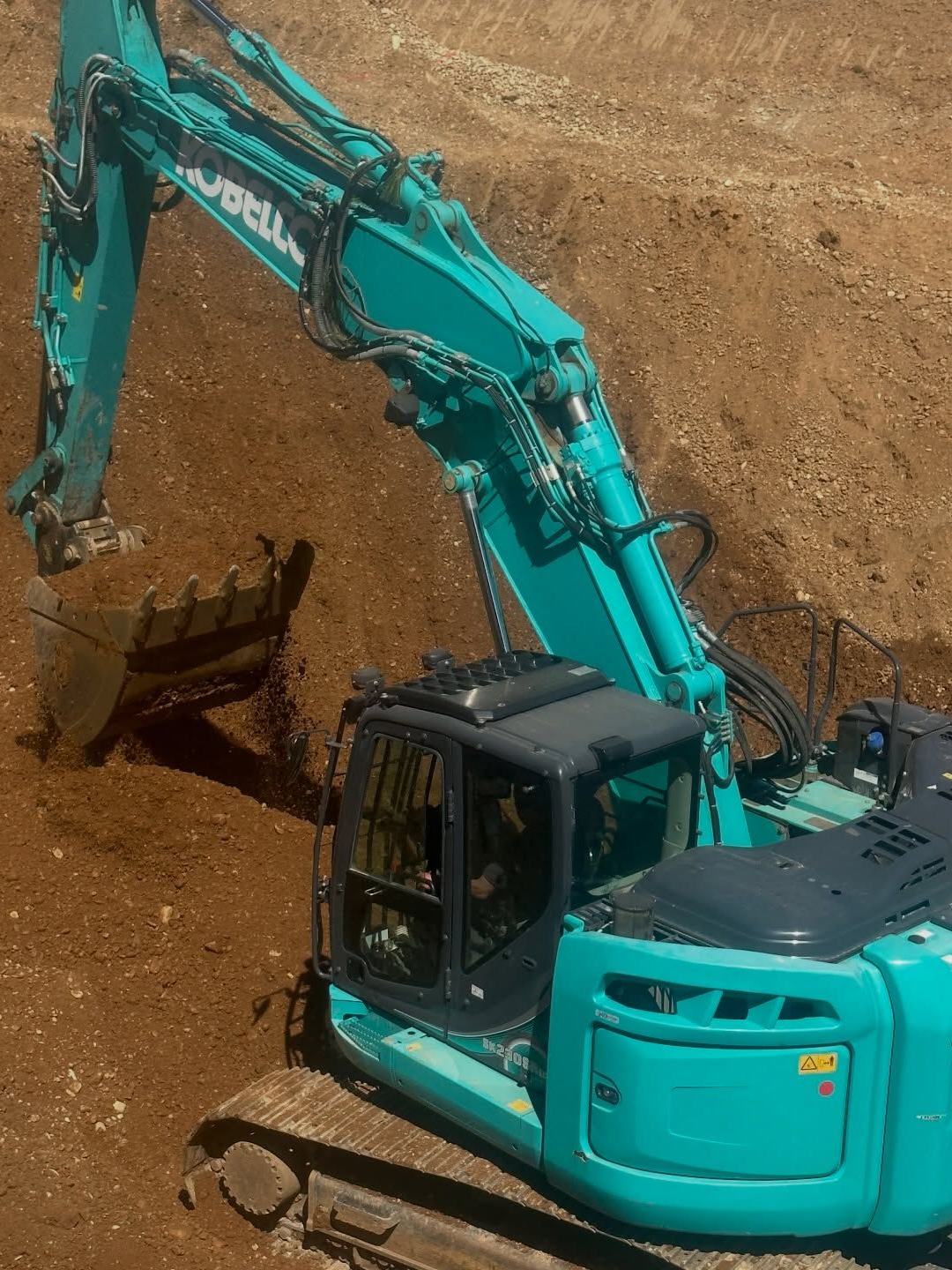 Une pelle mécanique turquoise creuse la terre sur un chantier, son godet levé au-dessus d'une pente de terre.