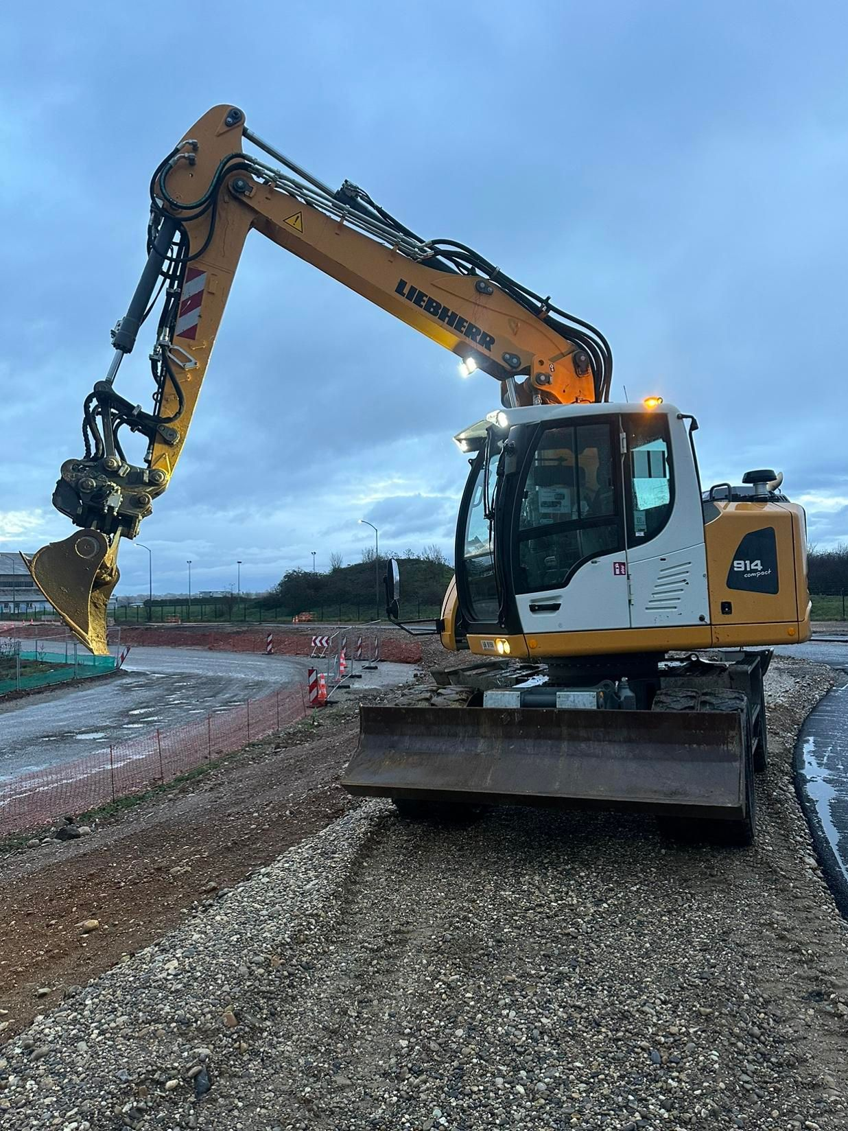 Une pelleteuse jaune sur un chantier de construction, au bord d'un chemin de terre humide sous un ciel nuageux.