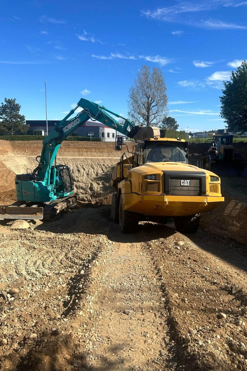 Une excavatrice charge de la terre dans un camion-benne jaune sur un chantier de construction sous un ciel bleu.