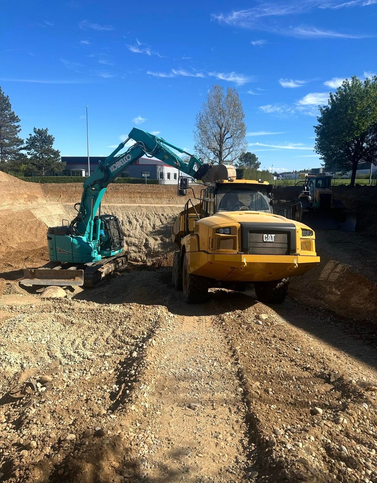 Une excavatrice et un camion-benne jaune sur un chantier de terrassement sous un ciel bleu.