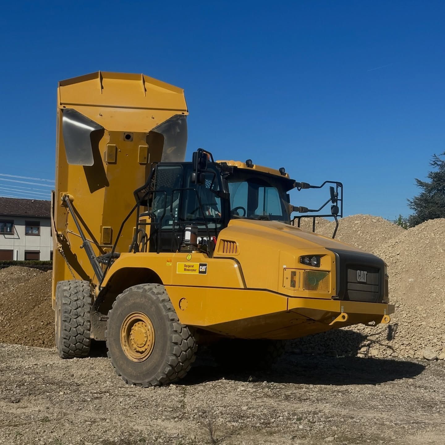 Un camion-benne jaune à plateau surélevé est stationné sur du gravier sous un ciel bleu azur.