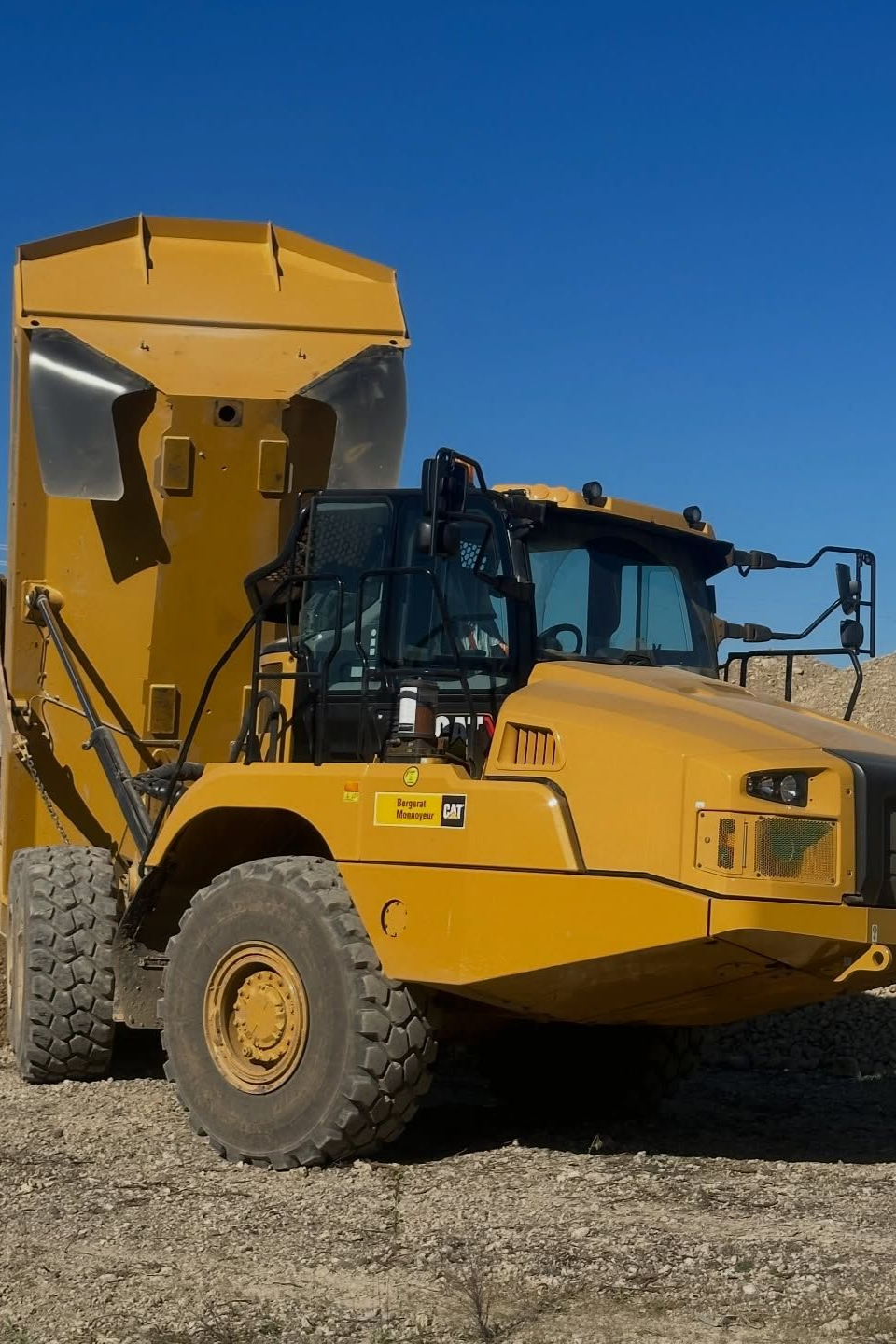 Camion-benne articulé jaune sur un terrain gravillonné sous un ciel bleu azur.