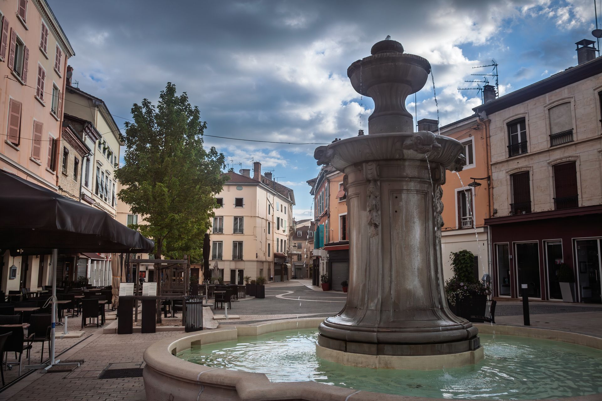 Place du village avec une fontaine en pierre, des terrasses de café et des bâtiments historiques sous un ciel nuageux