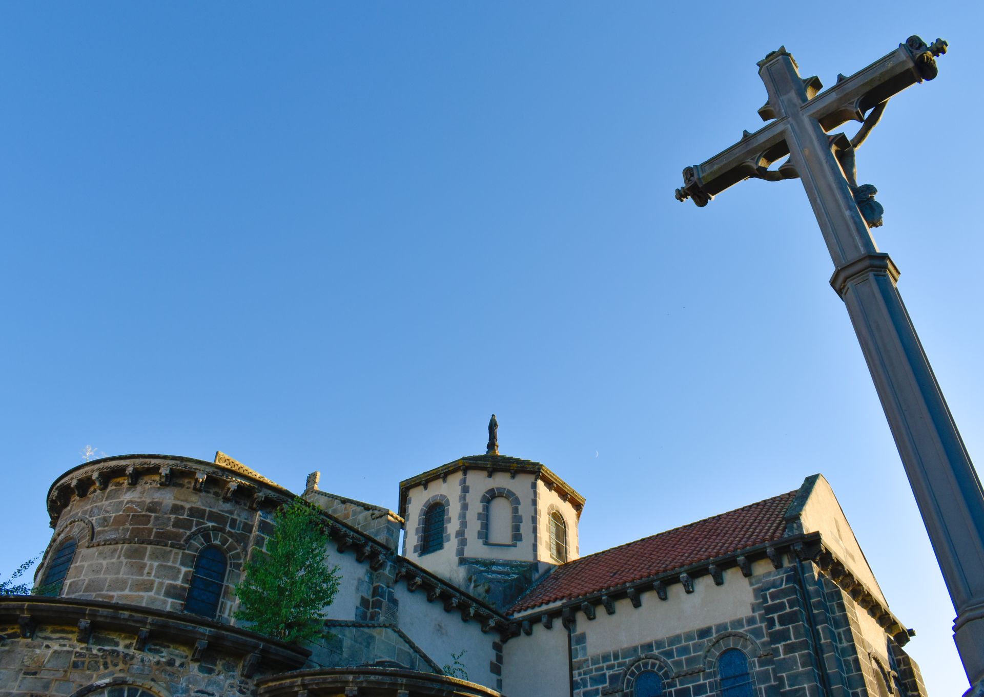 Église en pierre au toit rouge et au clocher, à côté d'une haute croix se détachant sur un ciel bleu.