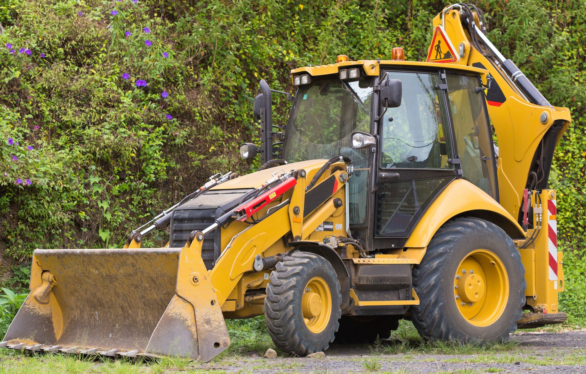 Une chargeuse-pelleteuse jaune est garée sur l'herbe à côté d'une haie feuillue.