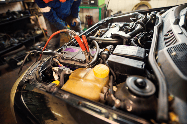 Compartimento del motor del coche con cables de arranque conectados a la batería, mecánico al fondo.