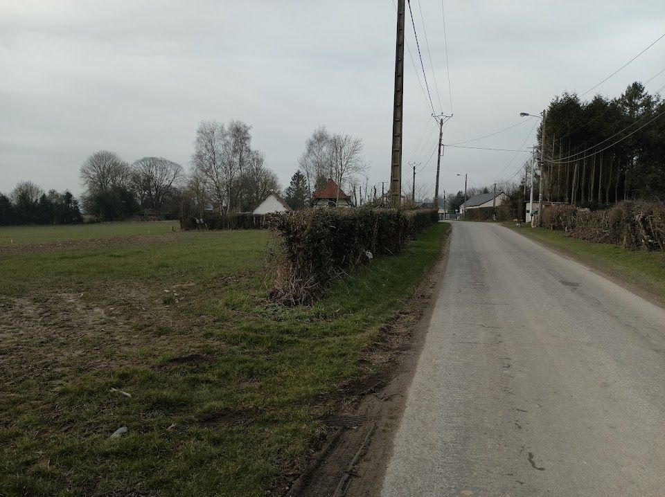 Route longeant un champ herbeux et des haies taillées, avec des arbres et un bâtiment en arrière-plan. Ciel nuageux.