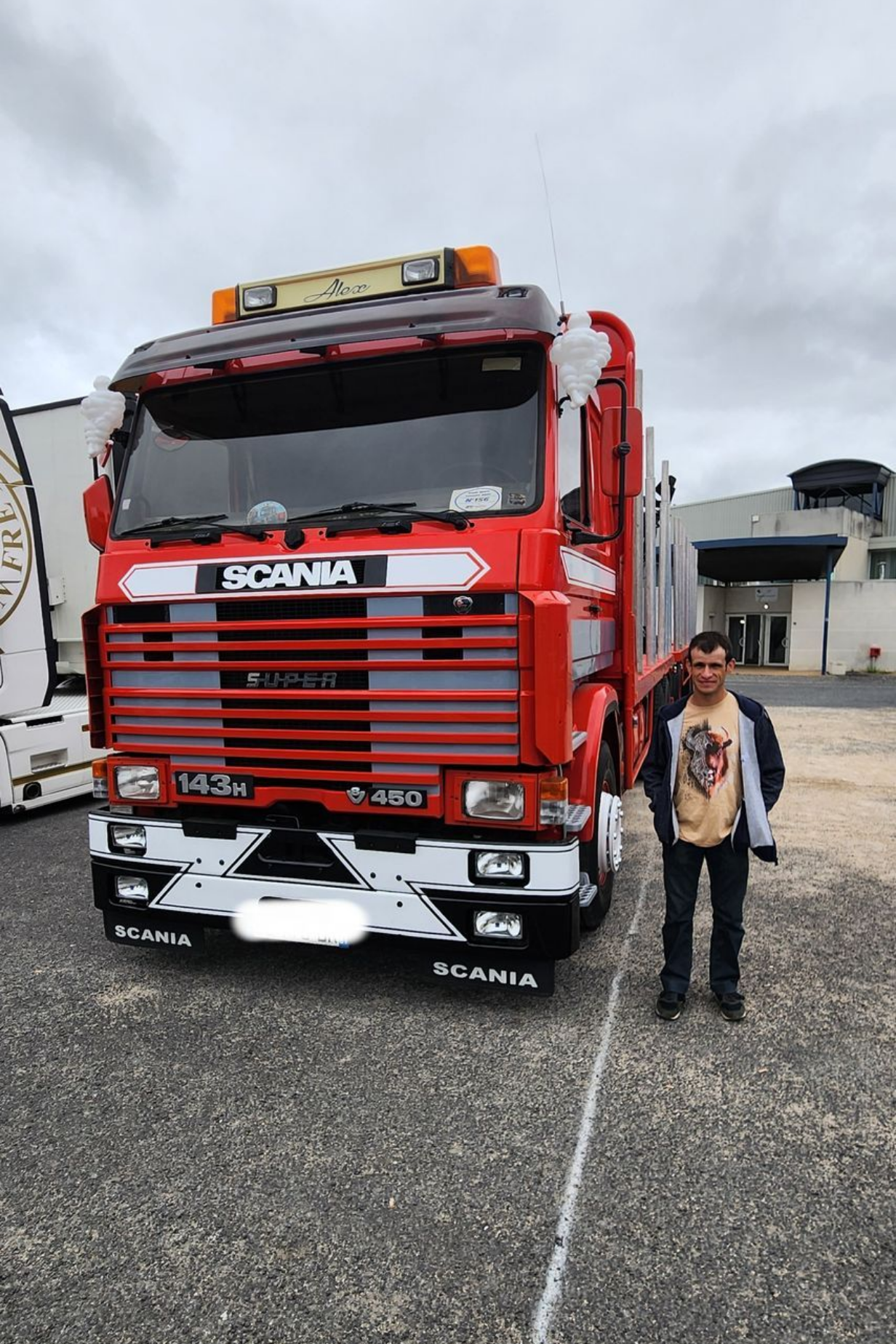 Un semi-remorque Scania rouge est stationné à l'extérieur, le gérant des réparation pose devant.