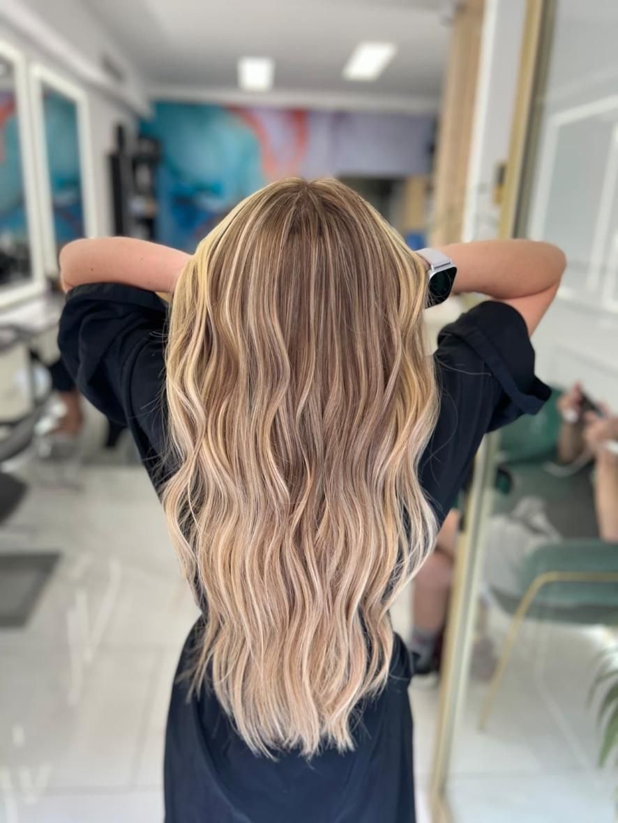 Persona con cabello rubio largo y ondulado vista de espaldas en una silla de peluquería, con los brazos levantados