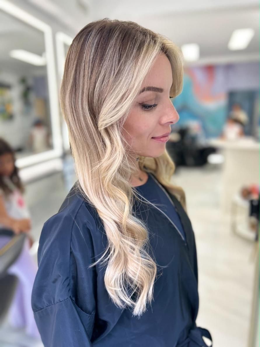 Vista de perfil de una mujer con largas ondas rubias en una peluquería, vestida con una blusa azul marino y sonriendo