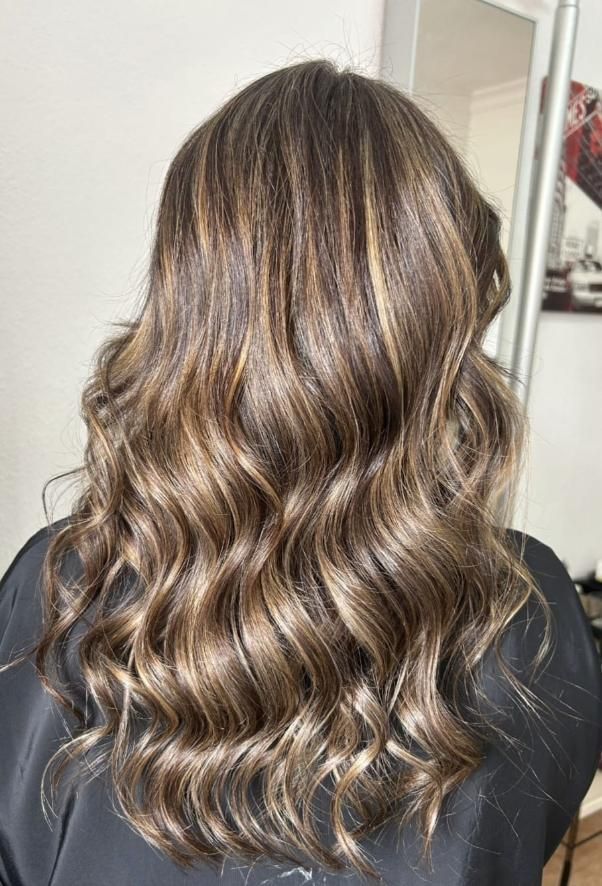 Cabello largo y ondulado castaño con reflejos rubios, peinado en rizos sueltos hacia atrás.