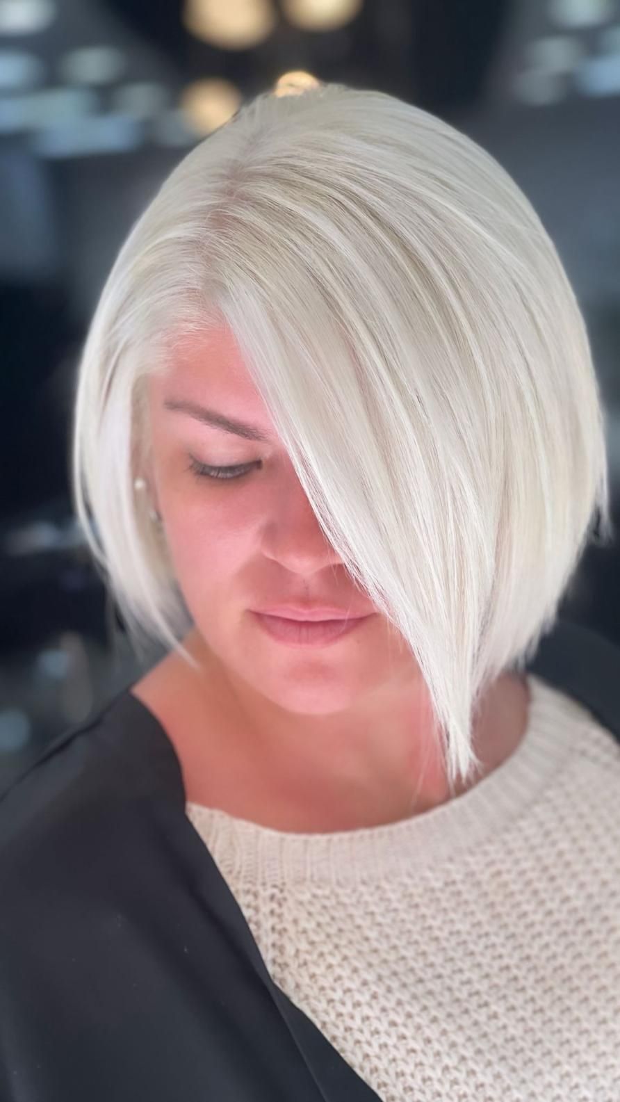 Persona con cabello corto rubio platino mirando hacia abajo, vistiendo un suéter claro sobre un fondo borroso.