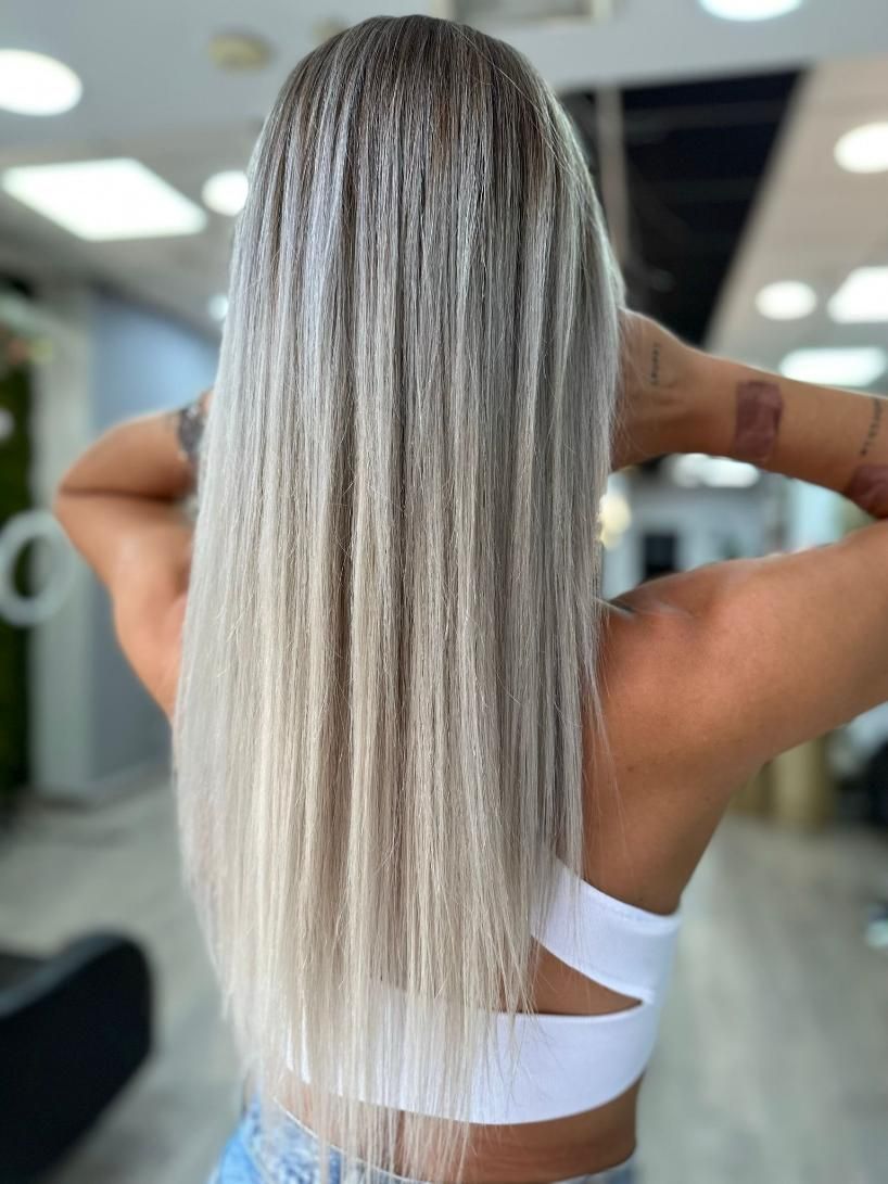 Persona con cabello largo rubio plateado de pie en una peluquería, vista desde atrás.