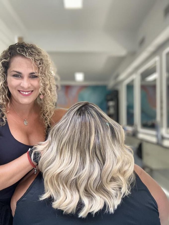 Estilista aplicando un tratamiento al cabello rubio ondulado de una clienta en el pasillo de un salón de belleza.