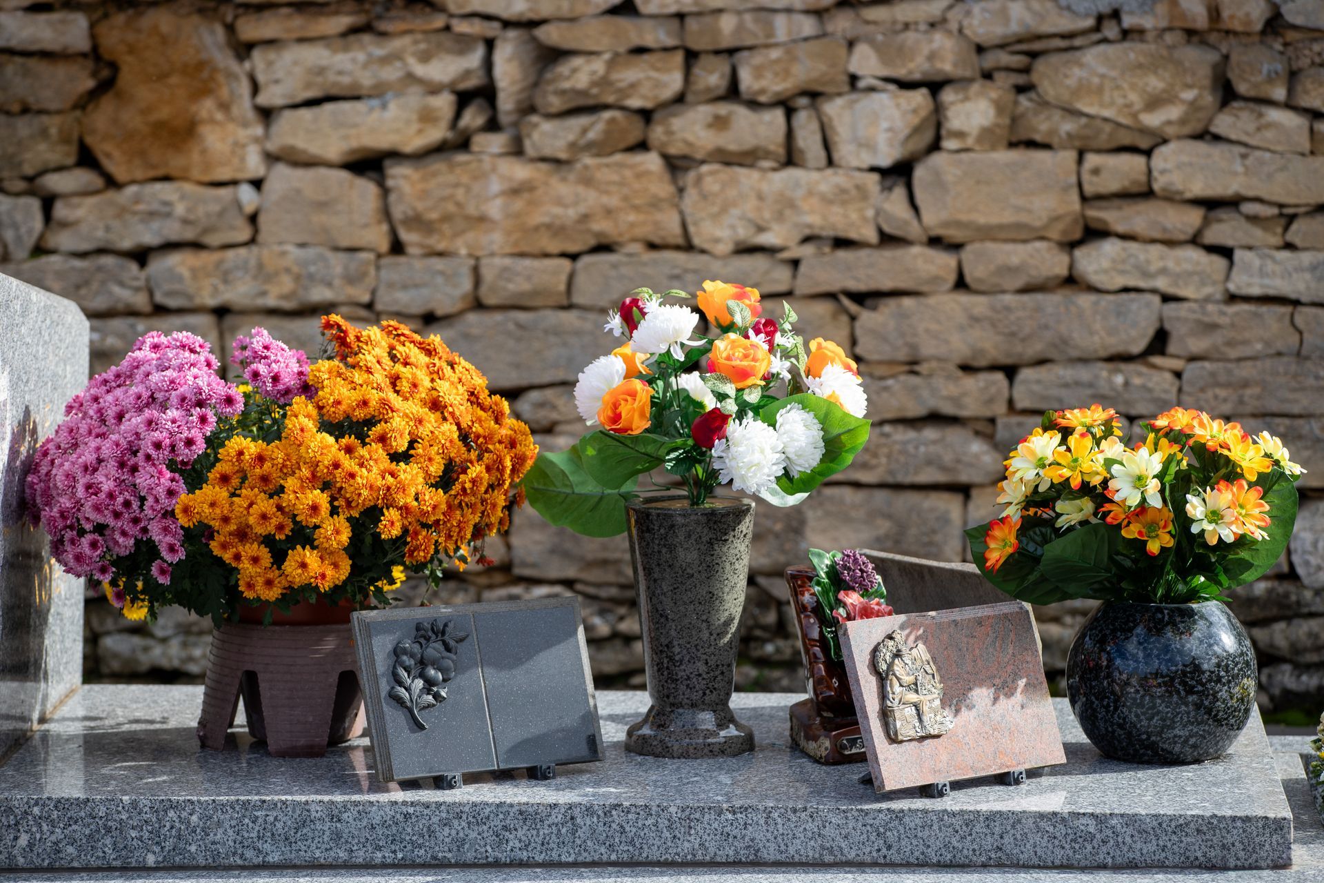 Des fleurs colorées dans des vases et des pots, disposées sur un rebord de pierre contre un mur de pierres empilées.
