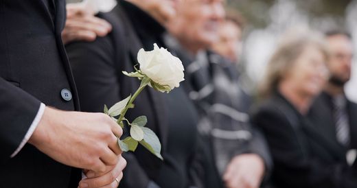 Une personne tient une rose blanche lors d'un enterrement, les personnes en deuil étant floutées en arrière-plan.