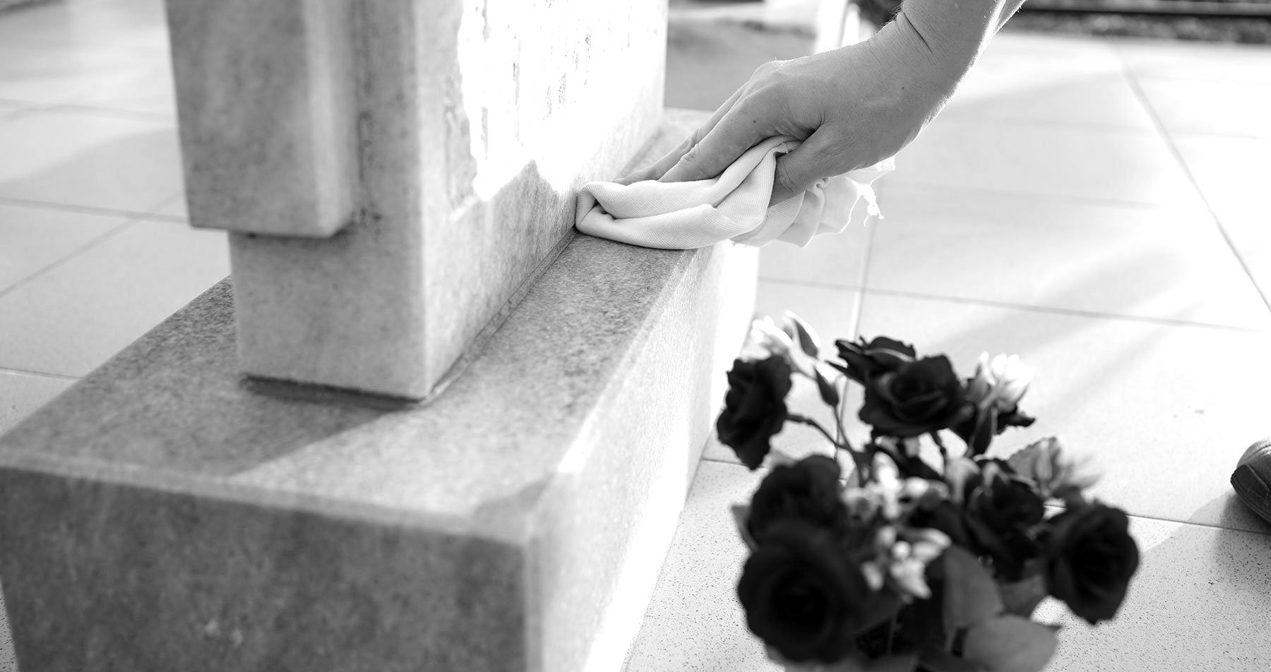 Une main nettoyant un monument de pierre à l'aide d'un chiffon, à côté de fleurs sombres sur un sol carrelé