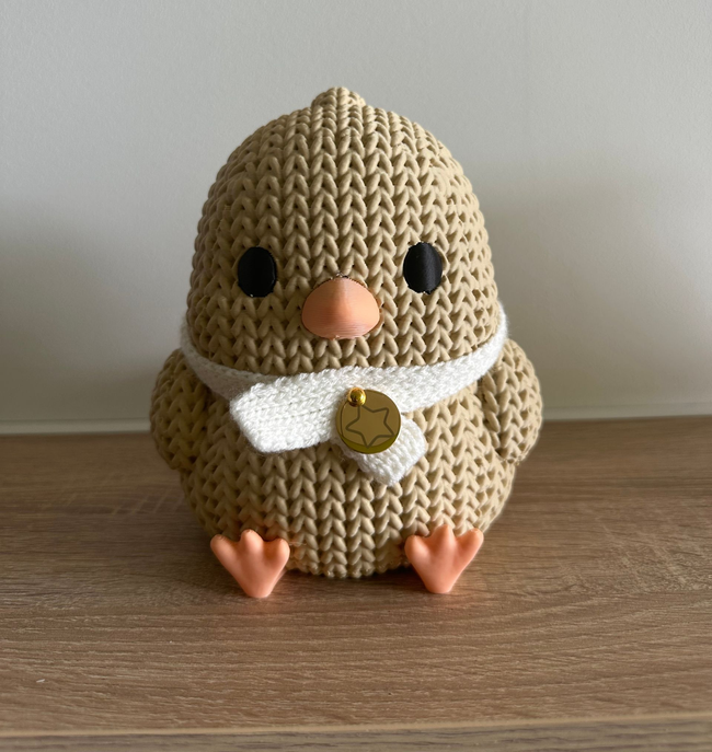 Poussin en peluche beige crocheté, aux yeux noirs, au bec et aux pattes couleur pêche, assis sur une étagère en bois avec une écharpe blanche.