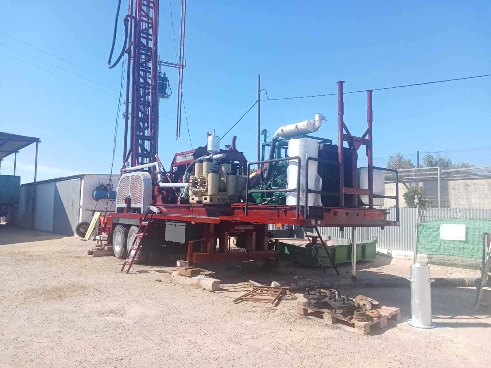 Plataforma de perforación industrial sobre una plataforma roja en un terreno polvoriento al aire libre bajo un cielo azul despejado.