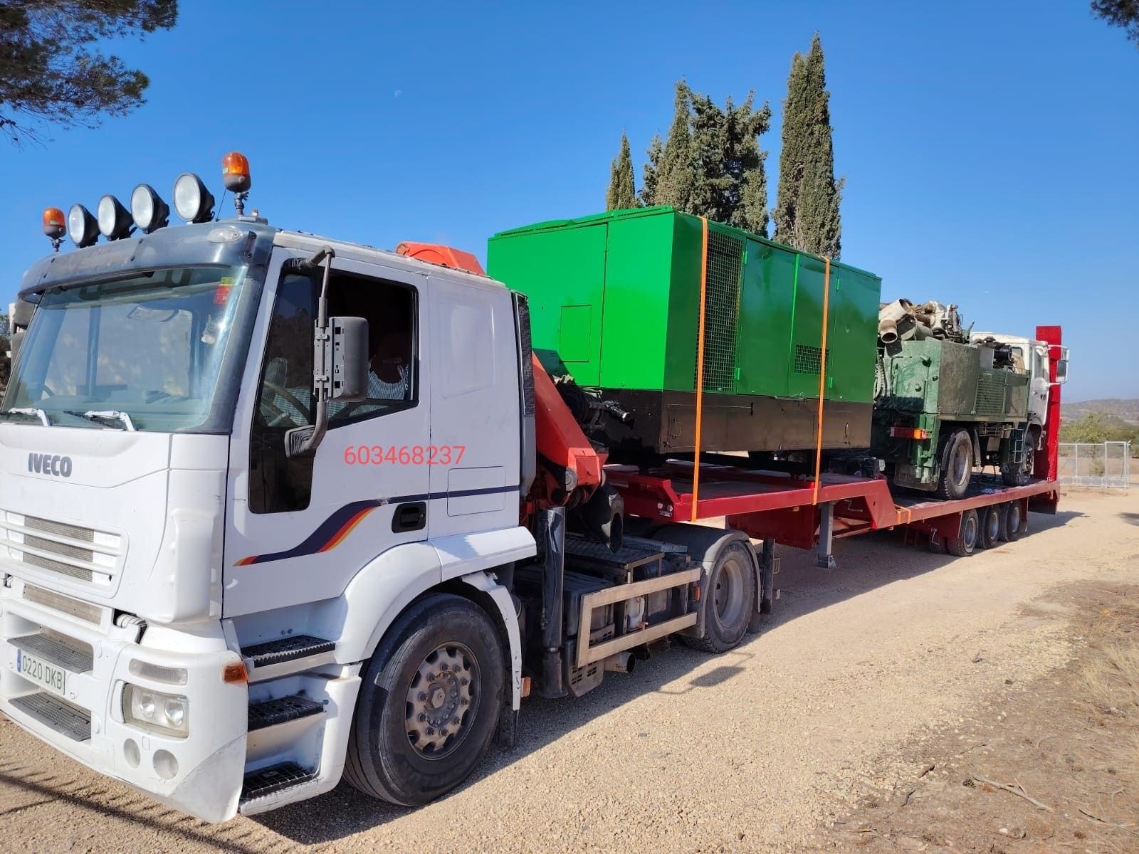 Camión blanco de plataforma plana transportando una gran máquina verde en un camino de tierra, con árboles al fondo.