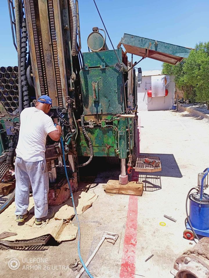Un trabajador se encuentra junto a una plataforma de perforación verde en una calle soleada, con mangueras y equipos cerca.