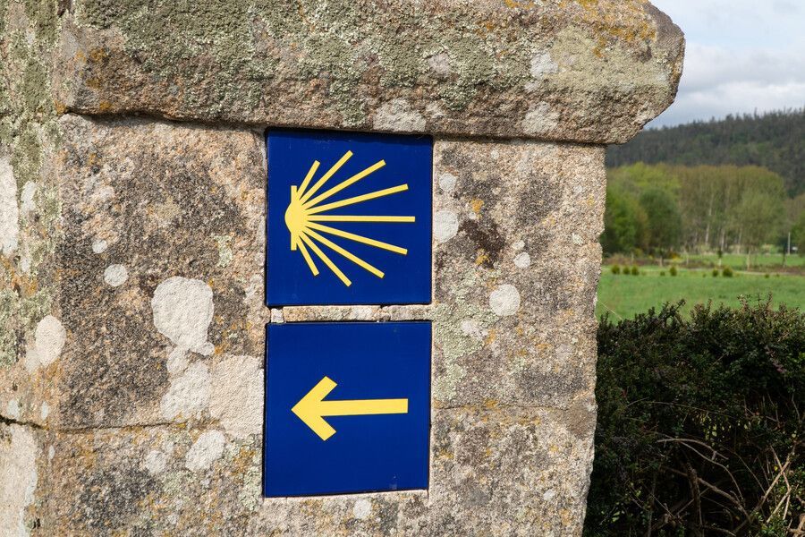 Señales azules del Camino de Santiago en un muro de piedra, con un símbolo de vieira amarilla y una flecha hacia la izquierda.