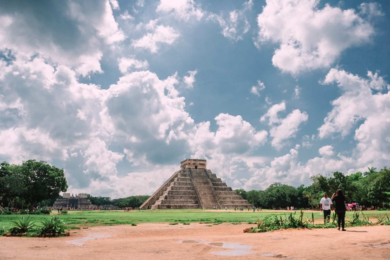 La pirámide de El Castillo en Chichén Itzá se alza sobre un campo de hierba bajo un cielo azul brillante y nublado.