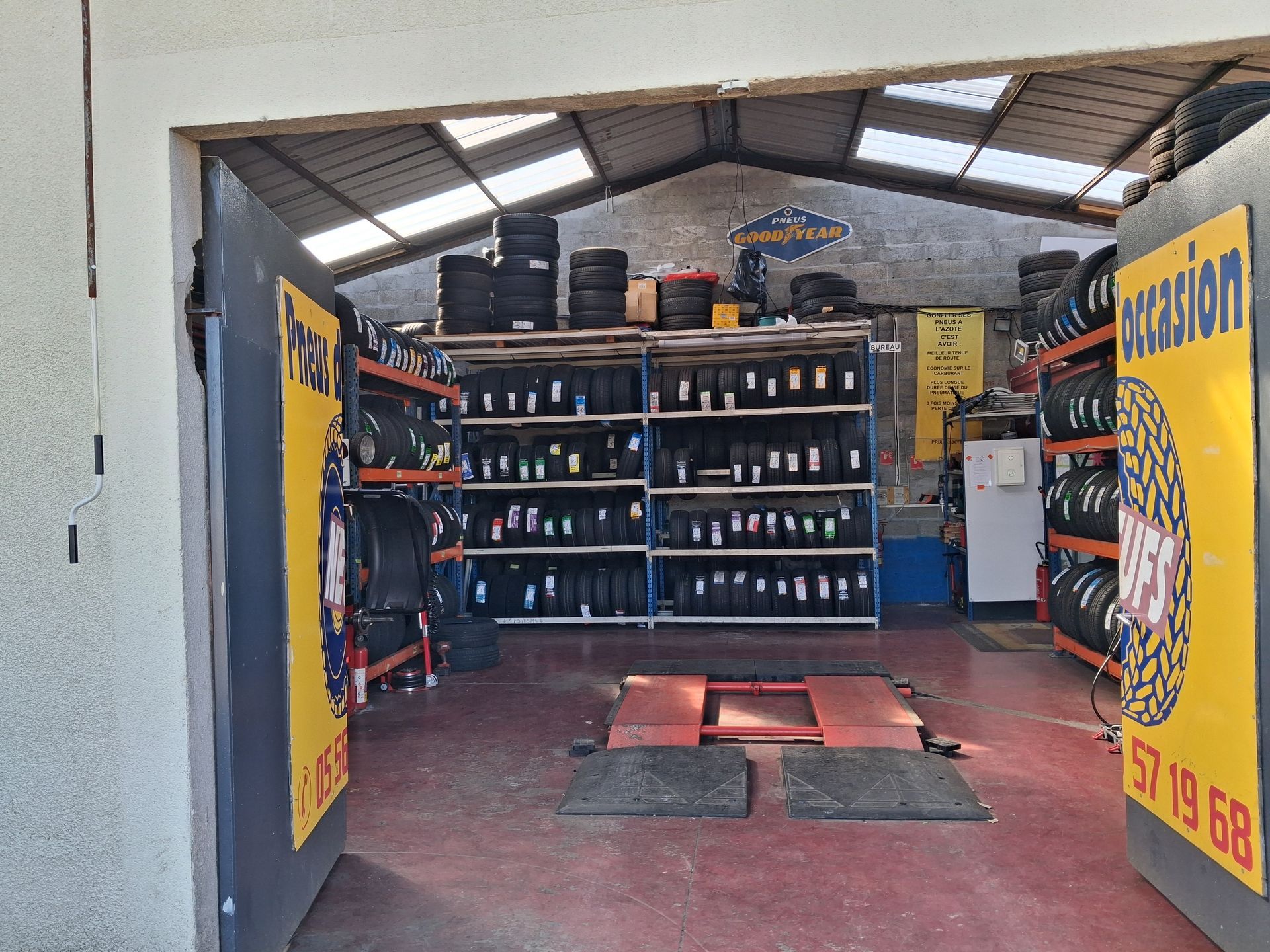 L'intérieur d'un garage automobile avec des piles de pneus sur des étagères métalliques, un pont élévateur hydraulique et de grandes enseignes près de l'entrée ouverte.