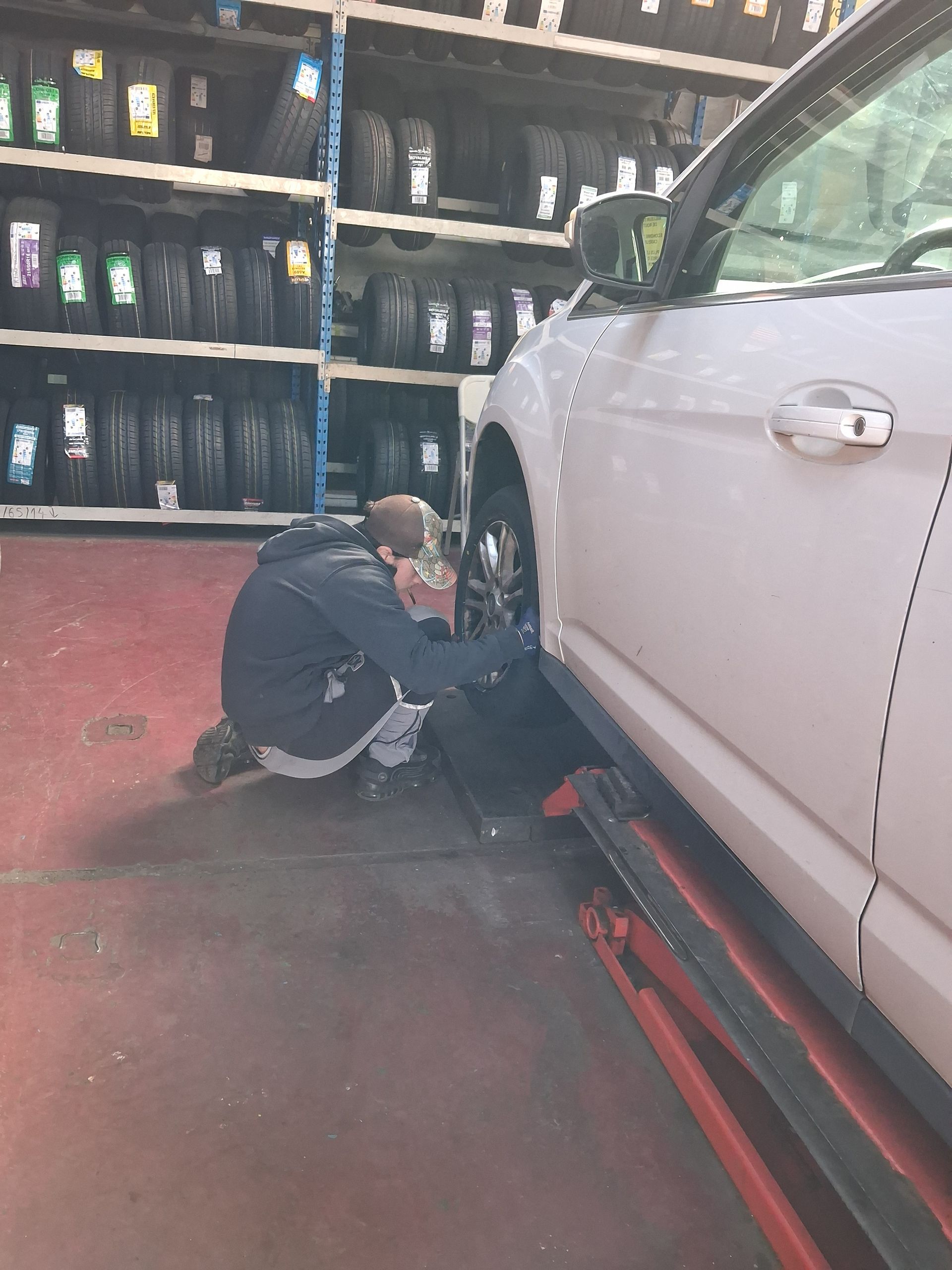 Un technicien s'agenouille pour réparer le pneu d'une voiture soulevée sur une plateforme hydraulique rouge à l'intérieur d'un atelier automobile équipé de supports à pneus.