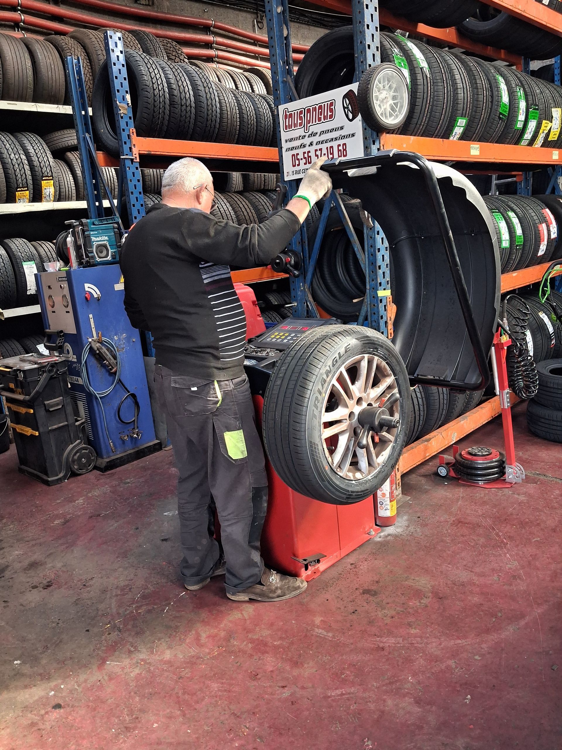 Une personne travaille dans un atelier de pneumatiques, en train d'équilibrer une roue de voiture sur une machine, avec des supports à pneus en arrière-plan.