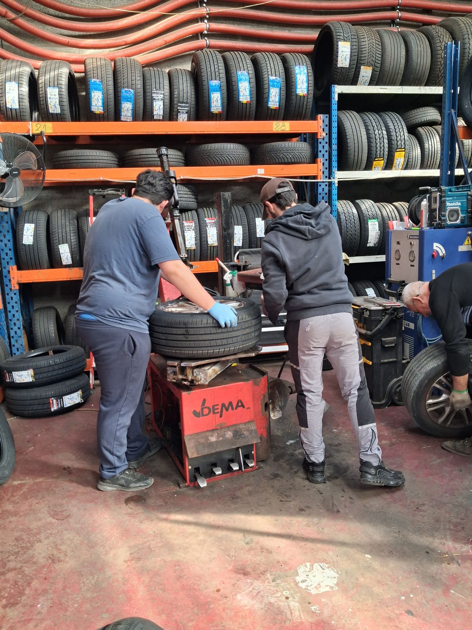 Deux personnes travaillent dans un atelier de pneumatiques, utilisant des machines pour monter et équilibrer des pneus, entourées d'étagères remplies de pneus en caoutchouc.